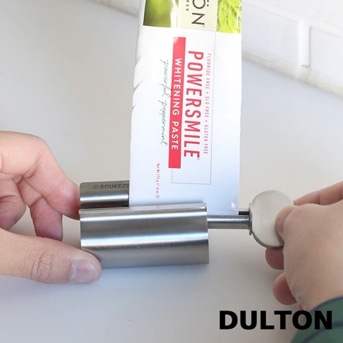 雙手正在操作一個 DULTON 金屬材質的牙膏擠壓器，不鏽鋼本體呈現筒狀結構，透過右側的轉柄旋轉，將一支標示著 POWERSMILE 的白色軟管牙膏捲入其中，以達到擠壓出剩餘牙膏的實用效果。
