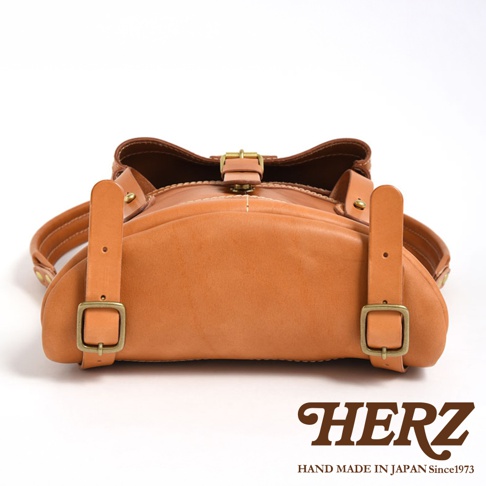 淺棕色皮革雙口袋折蓋後背包，頂部有兩個可調式皮帶扣，側面有兩個可調式皮帶和金屬扣。