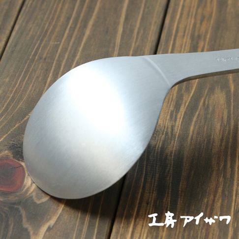 啞光銀色鋁製錘目紋湯勺，勺頭呈橢圓形，勺柄平直，置於深棕色木紋背景上。