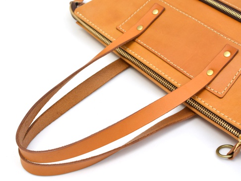 職人手作皮革威靈頓直式拉鍊托特包，呈現天然皮革溫潤的淺棕色調。包體結構清晰，帶有雙層皮革提把，以金屬鉚釘牢固固定。包身設有拉鍊開合，並點綴著精緻的縫線，展現細膩的工藝。