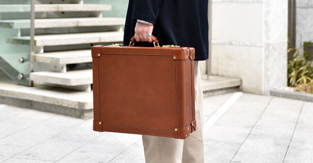 棕色皮革手提行李箱，配有金色金屬配件和提把，展示在戶外階梯背景前。