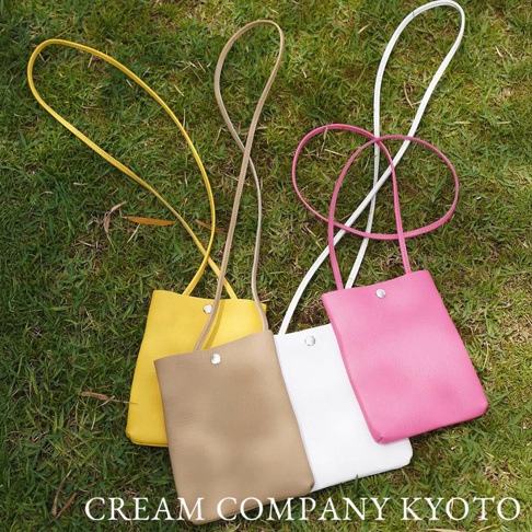 四款京都職人手作隨身手機包，顏色包括黃色、卡其色、白色與桃紅色，皆為皮革材質，附有細長背帶，並以單顆銀色金屬釦固定開口，散置於綠色草地上。