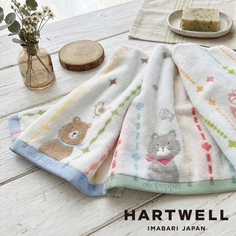 兩條印有可愛森林動物刺繡的 Hartwell 今治毛巾並排擺放在白色木質桌面上，左側毛巾飾有棕熊圖案與藍色邊框，右側毛巾則是灰色貓咪圖案配以綠色邊框，旁邊擺放著玻璃花瓶、木質杯墊及裝有肥皂的碟子，整體呈現溫馨清新的生活風格。
