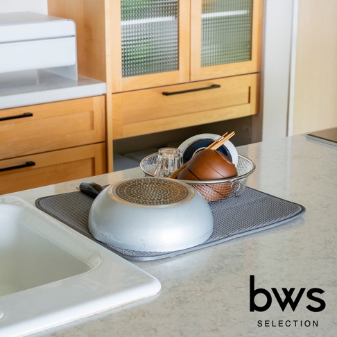灰色可折疊收納瀝水墊上放置著一個銀色平底鍋，鍋底朝上。旁邊有一個金屬網籃，籃中裝有玻璃杯、木製碗和筷子。