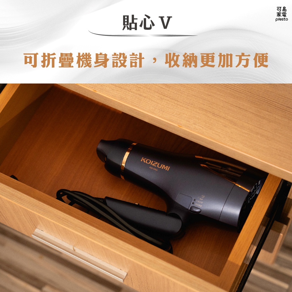 深灰色 KOIZUMI 負離子吹風機，帶有金色裝飾，放置於淺木色抽屜內，機身可折疊，方便收納。