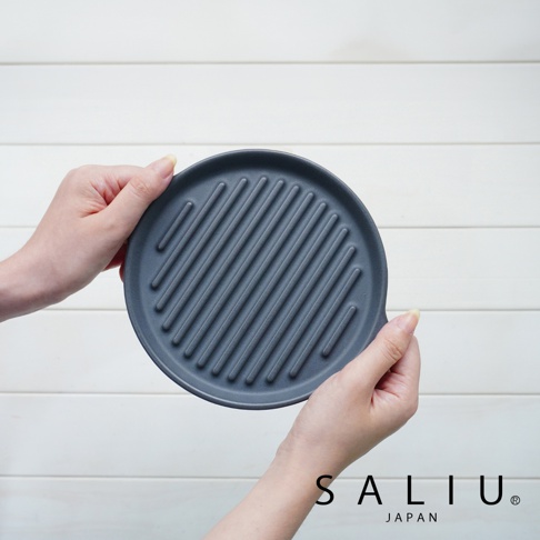 The chef 多功能耐熱陶製萬用波浪圓烤盤 S，為深灰色圓形陶製烤盤，表面佈滿平行波浪紋路，由雙手持握，背景為白色木紋。