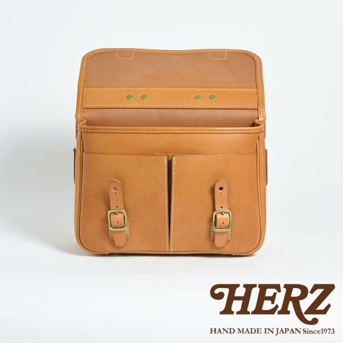 天然皮革製成的淺棕色兩用單肩包，正面有兩個帶有銅釦和皮帶的隔層，翻蓋內有三個黃銅鉚釘。