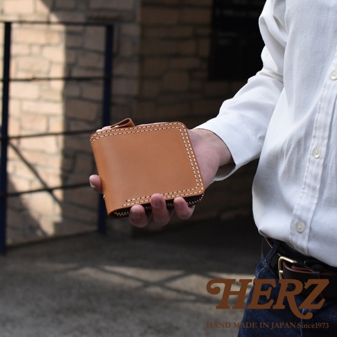 一位穿著白色襯衫的人手持一個淺棕色皮革製成的Herz職人手作全包式拉鍊短夾，皮夾表面呈現細緻的縫線工藝，畫面右下方印有品牌名稱與手工製造相關說明。