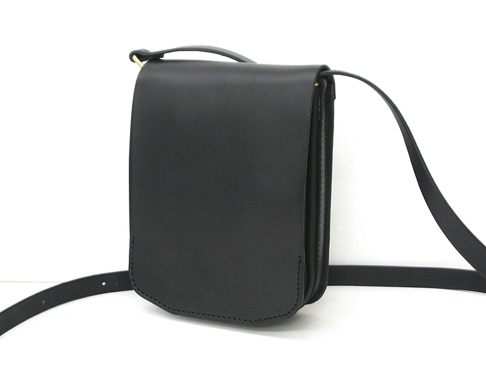 黑色皮革翻蓋直式單肩包，附有可調式肩帶和金色搭扣。包體線條簡潔，材質光滑，邊緣縫線工整。