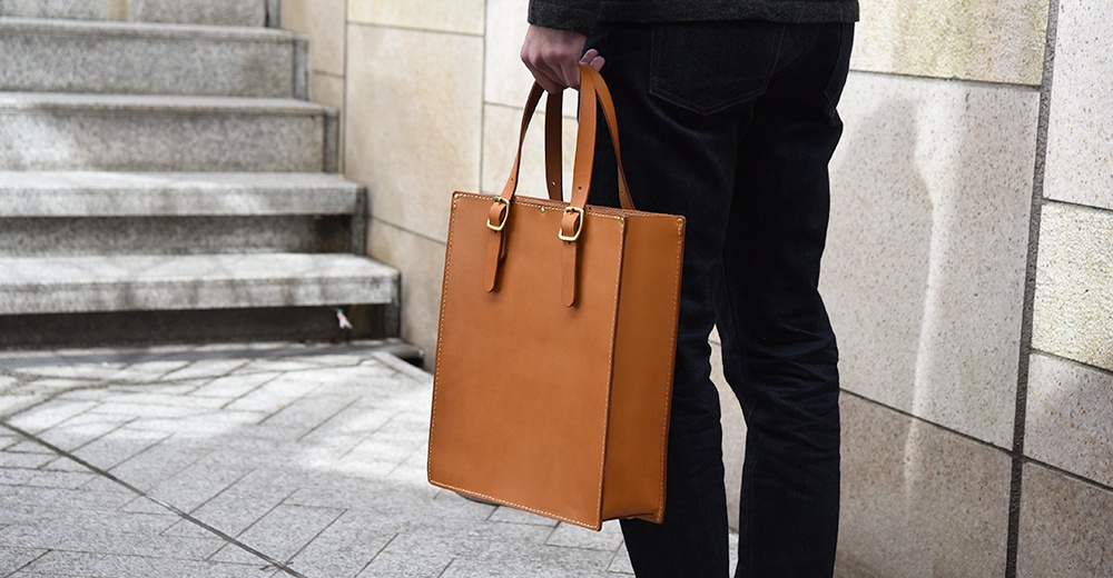 棕色皮革製成的托特包，有兩個提把和金屬扣環，由手持的男士展示。