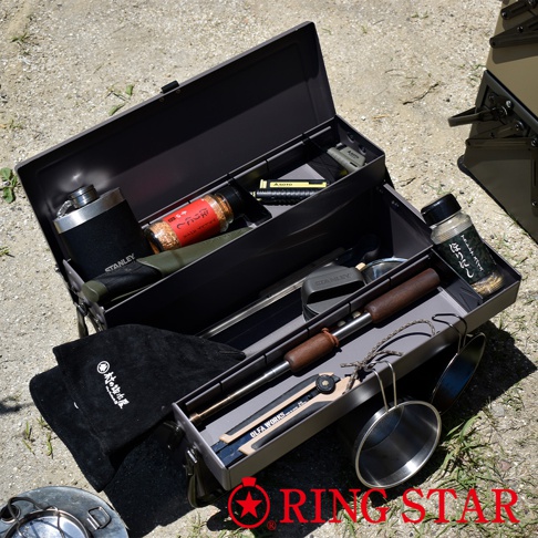 開放式灰色金屬工具箱，內含多樣露營或戶外用具，包括一個銀色保溫瓶、裝有紅色調粉末的玻璃瓶、一把綠色刀鞘的刀具、一個黑色燈具、以及帶有木質握柄的金屬烤叉。箱子下方還有一個黑色帆布袋和幾個金屬杯。