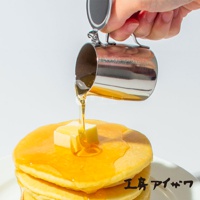 一個不鏽鋼附蓋蜂蜜盅，正將金黃色蜂蜜倒在一疊鬆餅和奶油上。