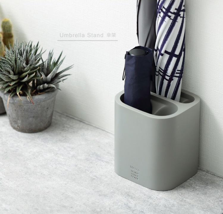 淺灰色簡潔雨傘筒架，側面有品牌標誌，可收納兩支雨傘，其中一支為深藍色摺疊傘，另一支為藍白條紋長柄傘。