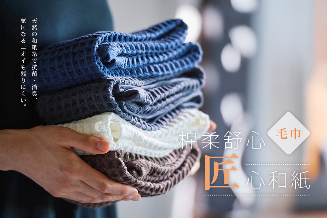 一手捧著疊成一疊的深藍、淺藍、白色和咖啡色毛巾，毛巾表面有網格紋理，左側有日文文字，右側有中文文字，標示著毛巾的材質和特性。