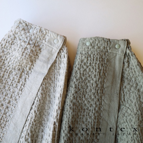 QULM 棉麻混紡平口鬆緊浴巾浴裙，展示米白色和淺綠色兩款。浴裙採用格子編織紋理，腰部有鬆緊帶設計，並以兩個淺色鈕扣固定。