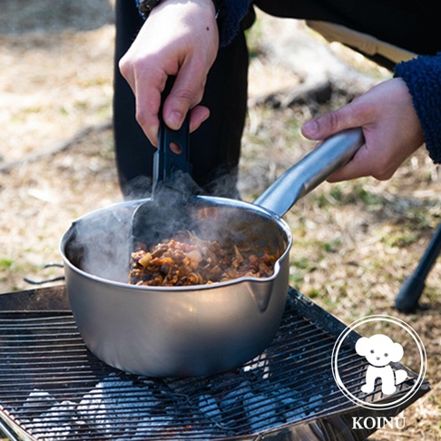 一隻手拿著黑色鍋鏟，在一個裝有食物並冒著蒸汽的銀色不鏽鋼雪平鍋中攪拌，鍋子置於烤架上，下方有燃燒的炭火。