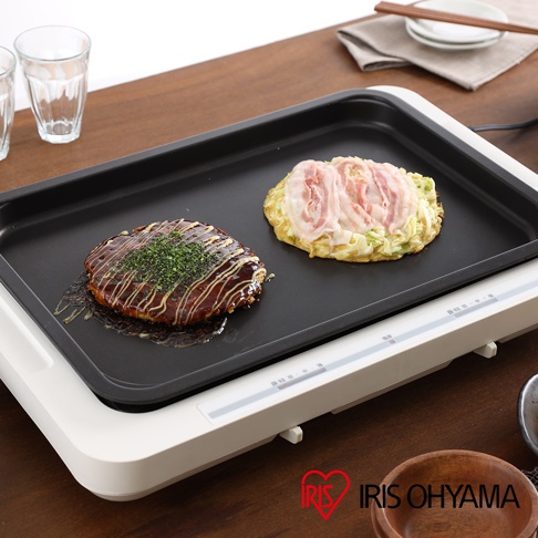 左右溫控電烤盤 WHP-012，黑色烤盤上烹調著兩份大阪燒，左邊一份淋有醬汁並撒上蔥花，右邊一份為豬肉片鋪在蔬菜上，烤盤邊緣為白色，帶有溫度控制按鈕。