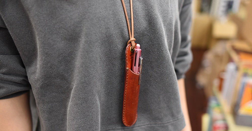 職人手作皮質掛繩筆套，內含一支粉色多功能筆，掛在深灰色上衣上，背景模糊。