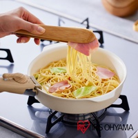 一個象牙白色的可拆卸陶瓷不沾平底鍋，鍋內盛有義大利麵、培根和高麗菜，正被竹製夾子夾起，鍋柄為木紋色，平底鍋放置於瓦斯爐上。