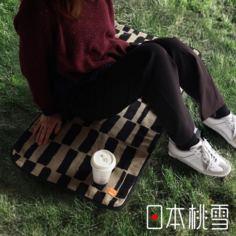 一個穿著深紅色毛衣和黑色褲子的人，坐在印有黑白長方形格紋圖案的毛巾上，旁邊放著一杯白色咖啡杯，以及帶有「日本桃雪」字樣的文字。