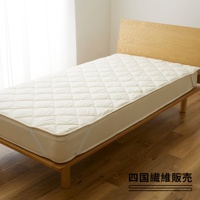 米白色菱格紋鋪棉床墊，邊緣有鬆緊帶，放置於木製床架上，背景為淺色牆壁與白色窗簾。