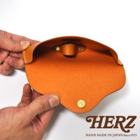 職人手作皮革眼鏡收納盒，橘棕色皮革，內部有固定帶與釦子，下方有品牌標誌HERZ HAND MADE IN JAPAN Since1973