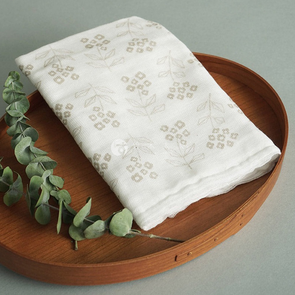 白色純棉花草紗布大拭巾，印有淺棕色小花圖案，疊放在木質托盤上，旁襯綠色尤加利樹枝。