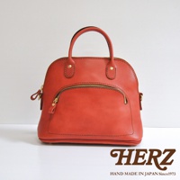 橘紅色皮革貝殼手提包，圓潤造型，設有雙提把、金色拉鍊及外層拉鍊口袋，風格經典，皮革質感細膩。