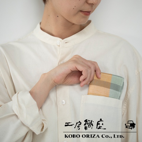 一名女子將一條橙色、灰色和綠色格紋的長方形手帕放入米白色襯衫的胸前口袋。手帕材質為織物。