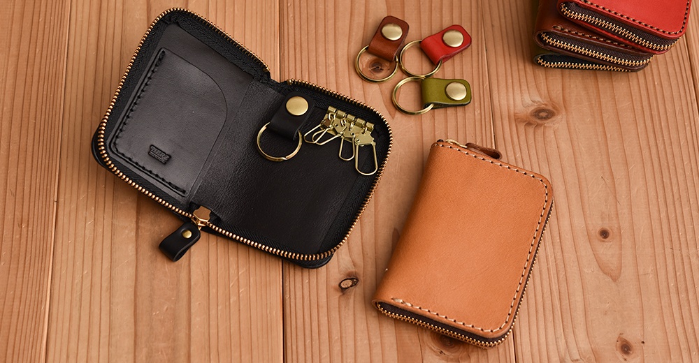 職人手作皮革多用途收納鑰匙包，包含展開的黑色皮革鑰匙包，內有四個鑰匙扣和一個卡槽，以及一個閉合的棕褐色皮革鑰匙包。另有三個獨立的皮革鑰匙圈，顏色為紅、棕、綠。