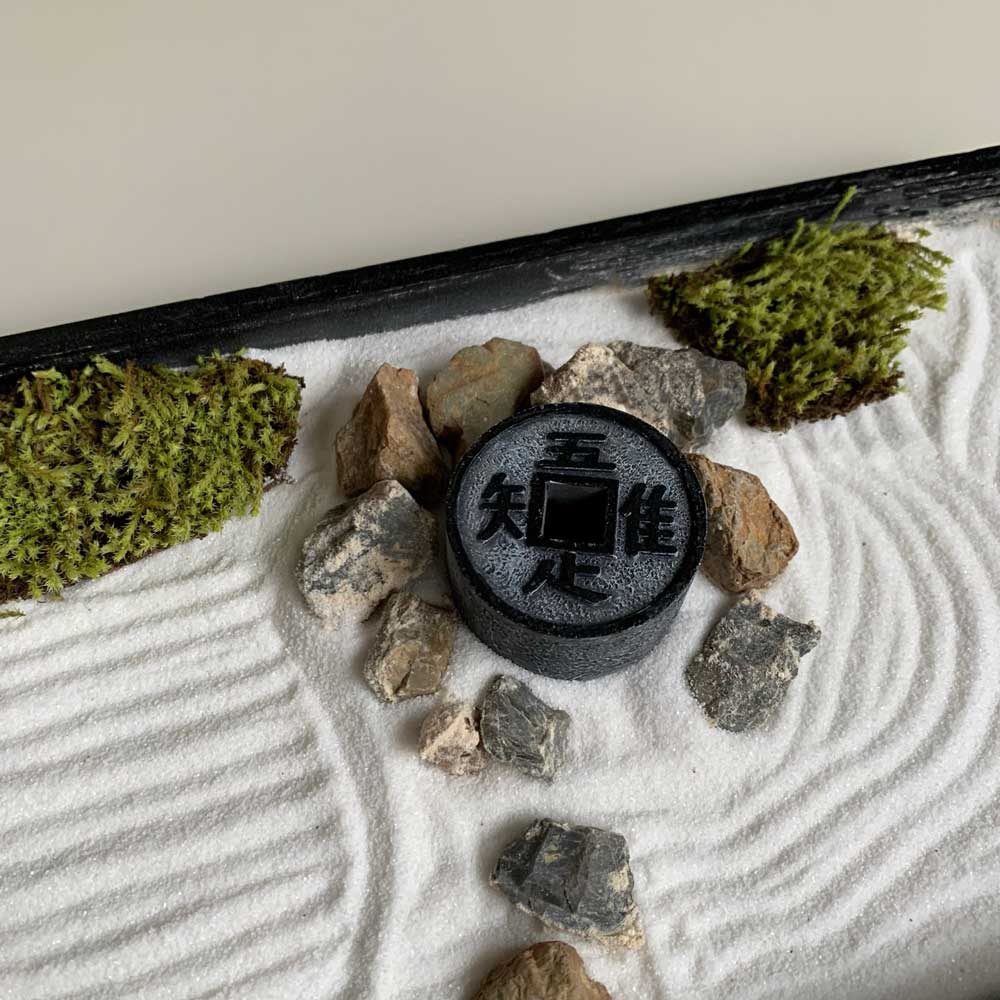 日本枯山水模型，白沙耙出細膩紋路，環繞著大小不一的淺灰色、棕褐色石頭，點綴著兩簇鮮綠的苔蘚。中央擺放一個黑色圓形模型，表面刻有漢字「知佳」，仿古銅錢造型。