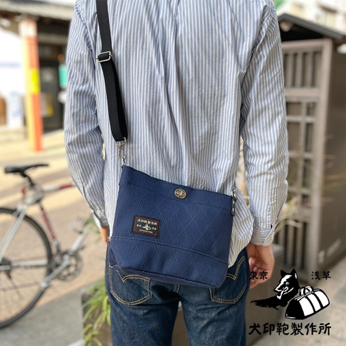 深藍色刺子織迷你側背包，斜挎於身穿條紋襯衫的模特身上，包身有菱格紋縫線和品牌標籤，搭配黑色可調式背帶。