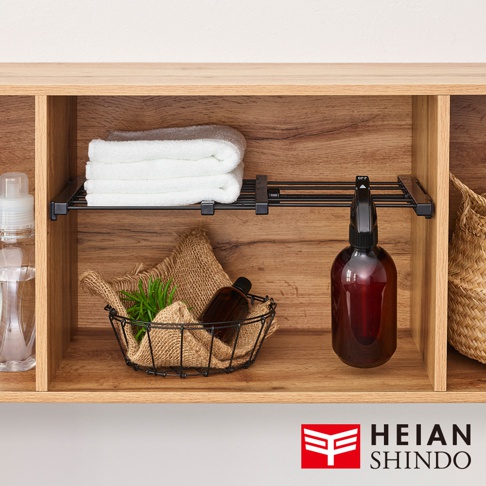 展示一個黑色的伸縮縫隙置物架，架上擺放著疊好的白色毛巾、一個裝有麻布和綠植的黑色線籃，以及一個琥珀色噴霧瓶。置物架安裝在木紋的層架空間內。