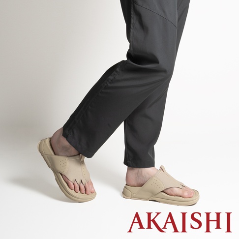 一位模特兒穿著卡其色的 AKAISHI 五指放鬆伸展拖鞋，搭配黑色長褲，從側面角度呈現，展示該鞋款的分趾設計與包覆性鞋面，背景為簡約的純白色。