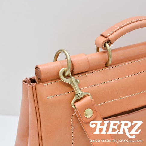 日本HERZ職人手作皮革屋瓦風側背包 CW-147，露出皮革的紋理與縫線細節，黃銅色的金屬掛鉤和旋鈕，包身上印有HERZ品牌標誌及「HAND MADE IN JAPAN Since1973」字樣