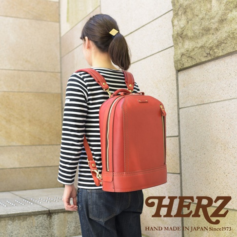 一名女性背著一個紅棕色皮革立體商務後背包，背包側面有拉鍊設計，肩帶和提把皆為皮革材質，搭配金屬扣環，整體風格簡約。