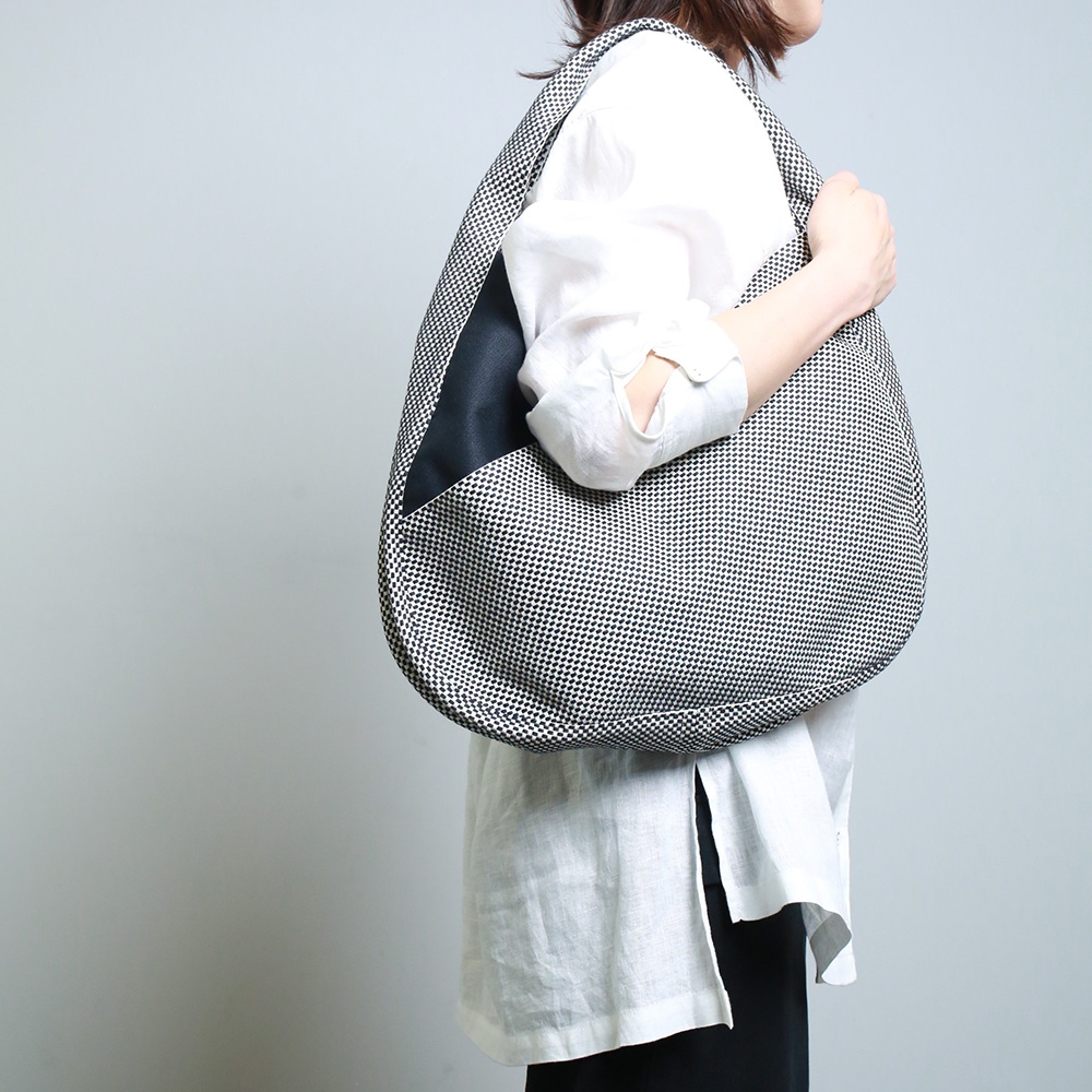 一名女子身著白色長袖襯衫，斜挎一個由黑白棋盤格紋刺子織面料製成的三河木綿半月包，包身一側有黑色皮革拼接。