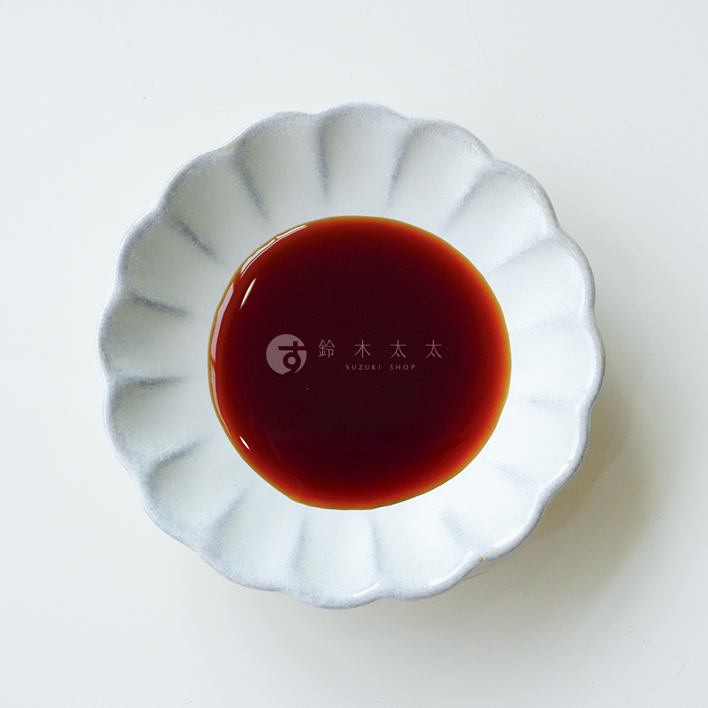 一個淺灰色的花瓣造型陶瓷碟，盛裝著深褐色、色澤飽滿的廣島牡蠣醬油，碟子中央印有 Suzuki Shop 鈴木太太的品牌商標，整體以俯視視角呈現，背景為乾淨簡潔的白色。