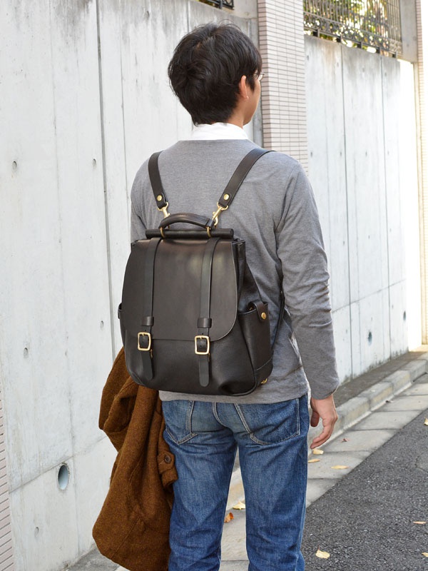 黑色皮革雙扣後背包，配有金色金屬配件和頂部提把，背負於身穿灰色長袖上衣和藍色牛仔褲的男性身上。