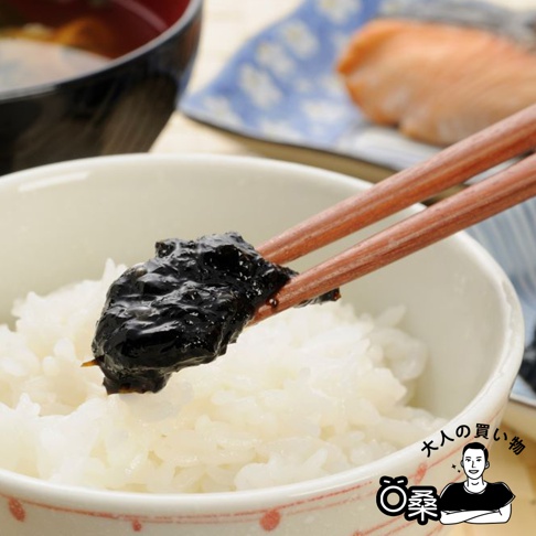 一雙木質筷子夾著一團深黑色的海苔醬，放在一碗白飯上。碗的邊緣有紅色圖案。背景中還有一個裝有湯的碗和一個裝有鮭魚的盤子。