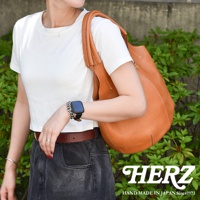 一位女性身穿白色短袖上衣，肩掛一個 tan 色皮革製成的圓形托特包。包包採用手工縫製，展現職人質感。