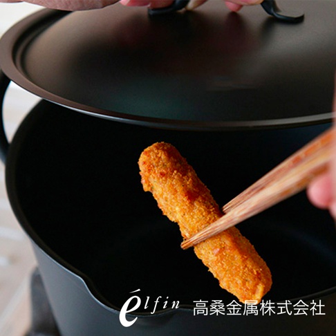 日製深型鍛鐵炸鍋，黑色鍋身，鍋蓋部分開啟，一根炸物被筷子夾起，準備放入鍋中。