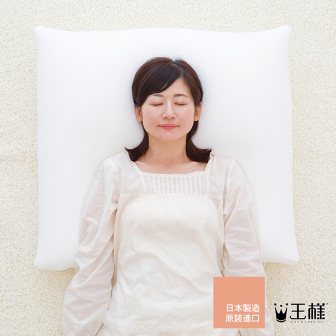 一位女性仰躺在純白色、材質柔軟的枕頭上，臉部朝上，閉著眼睛，頭髮為深棕色。她身穿寬鬆的淺色長袖上衣。枕頭放置在米白色、絨毛感的地毯上。右下角有一個橘色方形標籤，上面有日文文字和「王樣」品牌標誌。