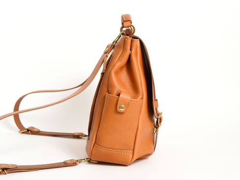 棕褐色皮革雙扣後背包，側面有方形口袋，配有可調式細肩帶和頂部提把，整體呈現簡約風格。