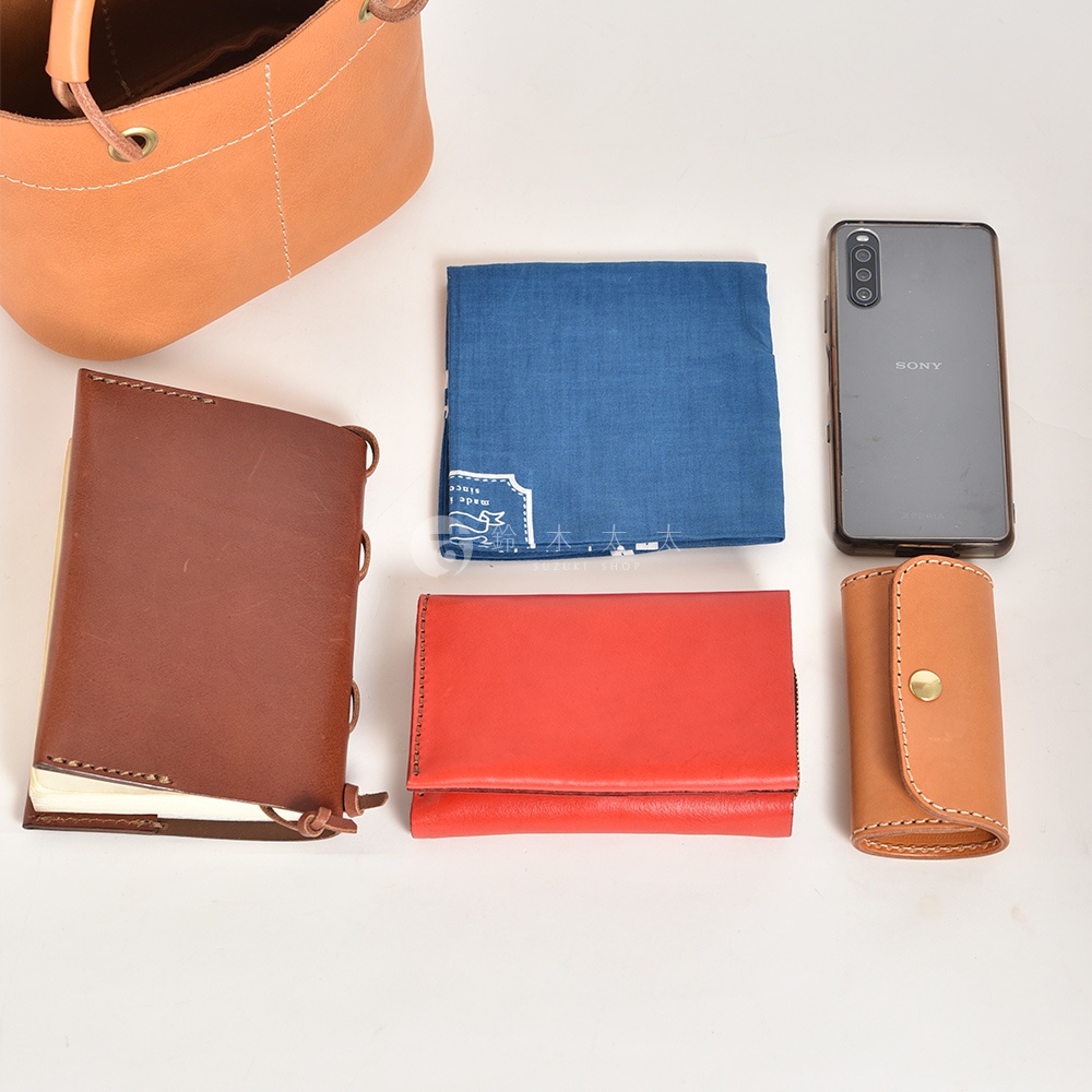 皮革製提把午餐袋，搭配棕色筆記本、紅色長夾、藍色印花手帕和手機。
