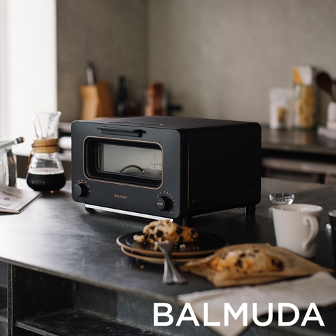 黑色 BALMUDA 蒸氣烤麵包機，配有金色邊飾，前方有玻璃門和兩個旋鈕。烤麵包機擺放在有咖啡器具和烤麵包的廚房檯面上。