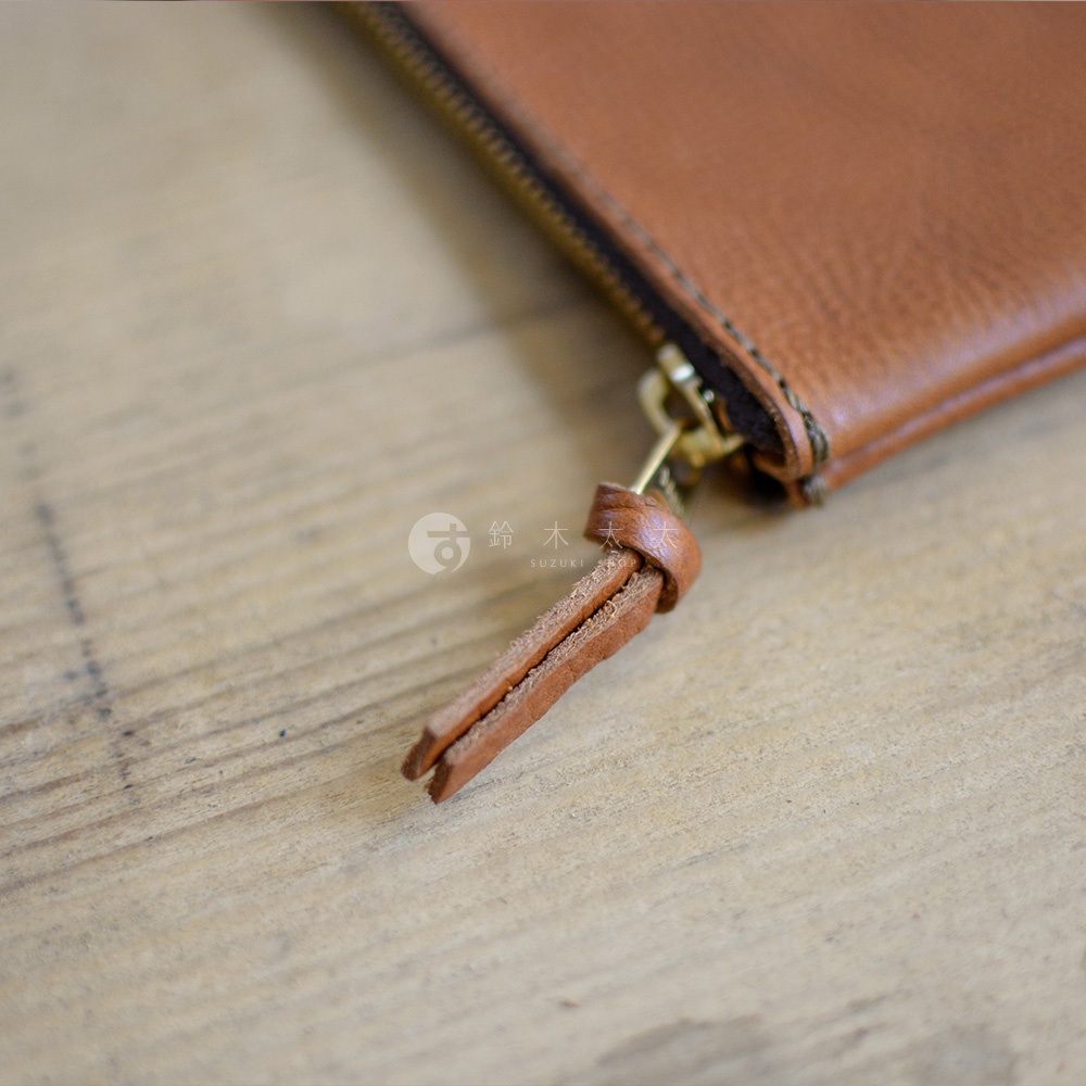 職人手作皮革Organ拉鍊式小物收納袋，特寫金色拉鍊與皮革拉環，收納袋為棕褐色皮革材質，置於木紋背景上。