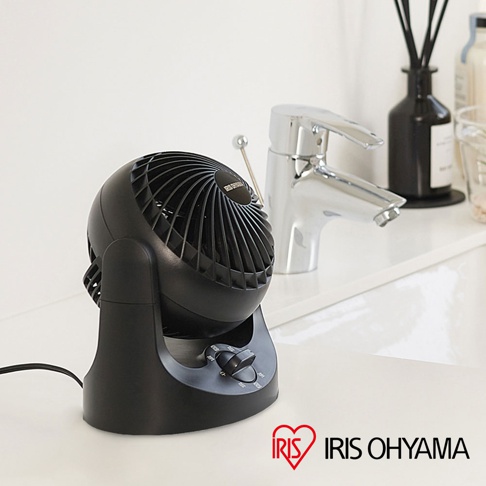 黑色桌上型空氣循環扇，具有可調整角度的球狀扇頭，底部有黑色旋鈕控制風速。產品擺放在白色檯面上，背景是鉻合金水龍頭和黑色擴香瓶。