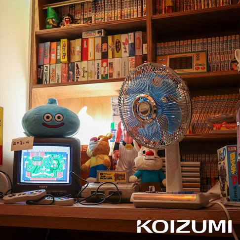 復古風格的白色電風扇，扇葉為藍色，帶有金屬網罩，放置在充滿懷舊遊戲機和商品的木質架子上。