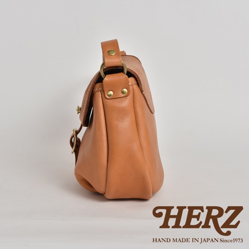 HERZ 職人手作皮革休閒旅行馬鞍包，側身視角，咖啡色皮革，經典馬鞍包造型，上方有提把和金屬釦，下方有品牌 LOGO。